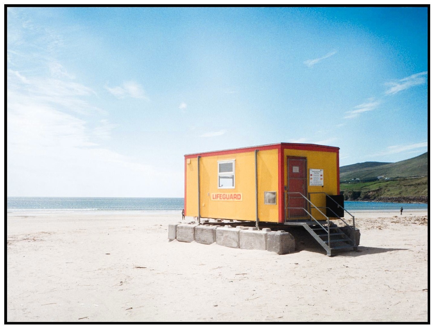 A yellow lifeguard hut with a red door stands on a sandy beach next to the ocean under a blue sky.