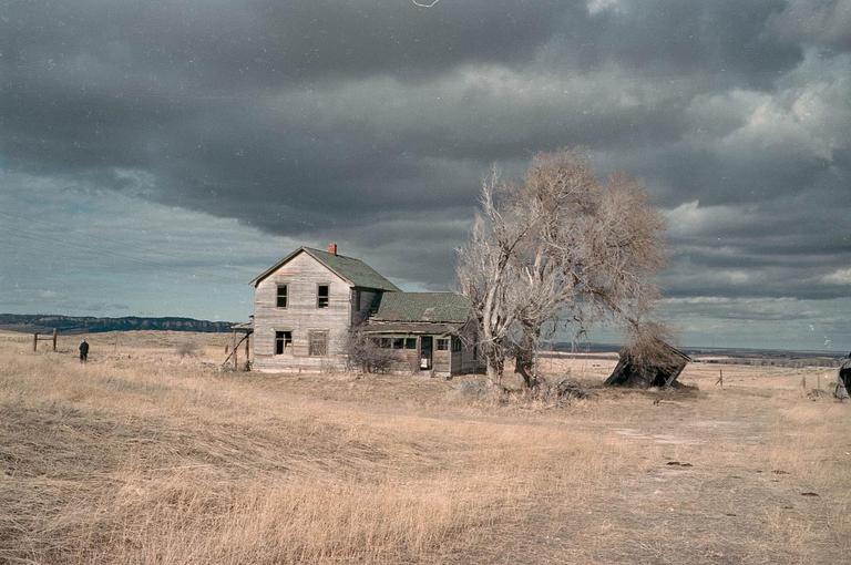 An abandoned house stands in a field under a dramatic cloudy sky, with a lone figure in the distance.