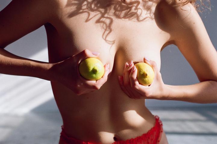 A woman covers her breasts with two lemons while standing in sunlight.