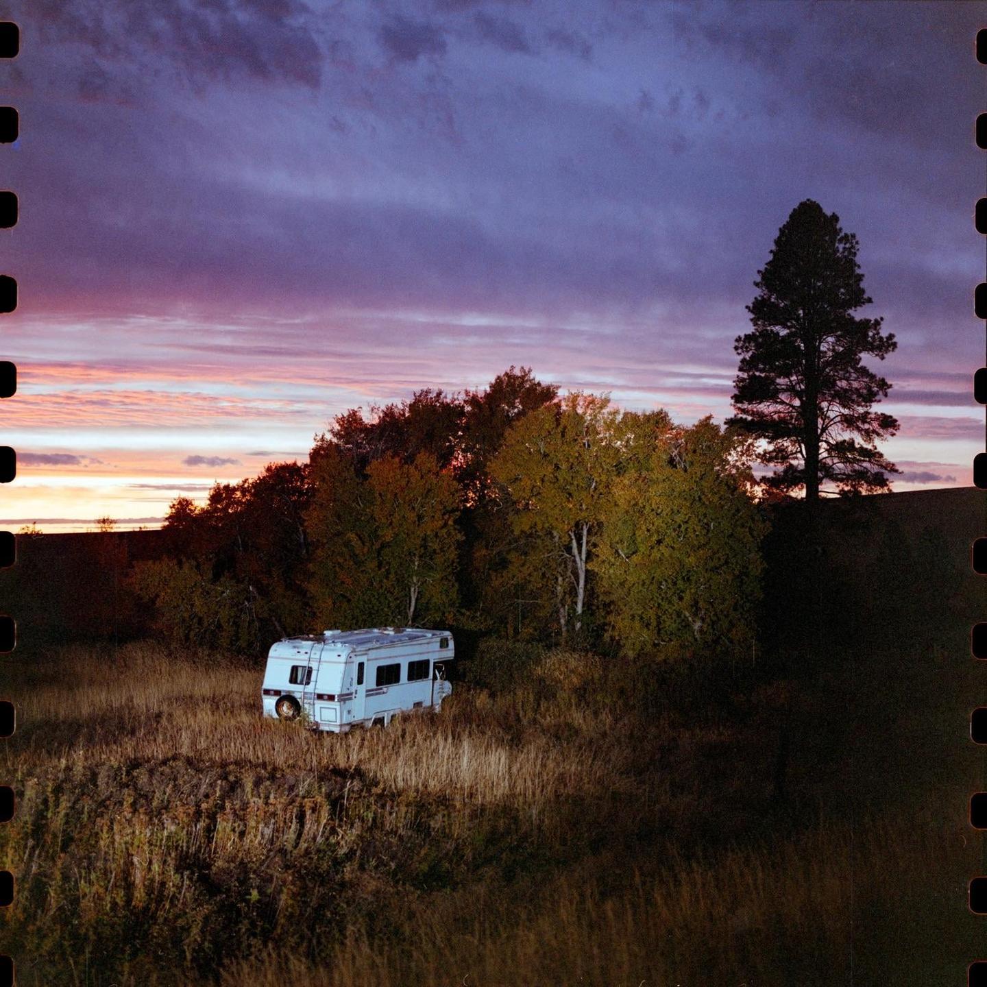 A white recreational vehicle is parked in a field with trees under a colorful sunset sky.