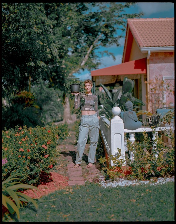 A woman stands on a brick path holding a potted plant in a lush garden next to a house with a red roof.