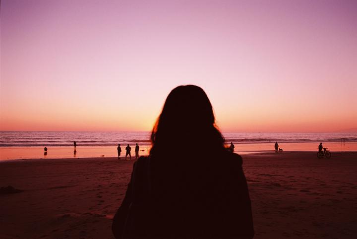 A person with long hair stands on a beach looking out at the ocean and sunset with other people in the distance.