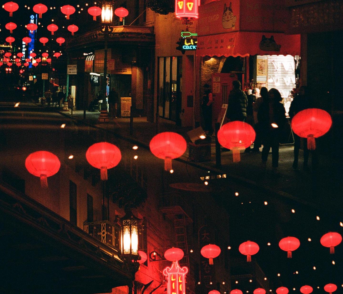 Red lanterns illuminate a street at night, with people walking past shops.