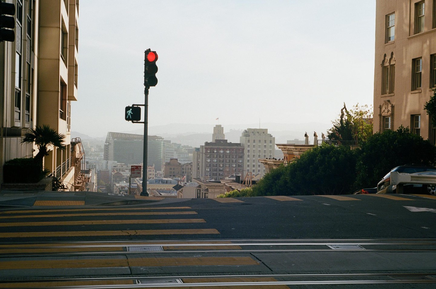 A red traffic light and a green pedestrian signal are visible on a steep street with buildings and a city in the background.