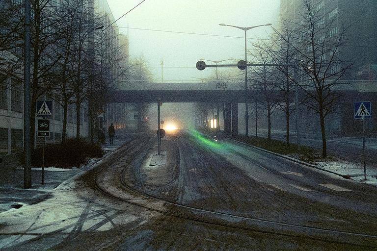 A street with tram tracks and bare trees is covered in snow and fog, with a bright light visible in the distance under a bridge.