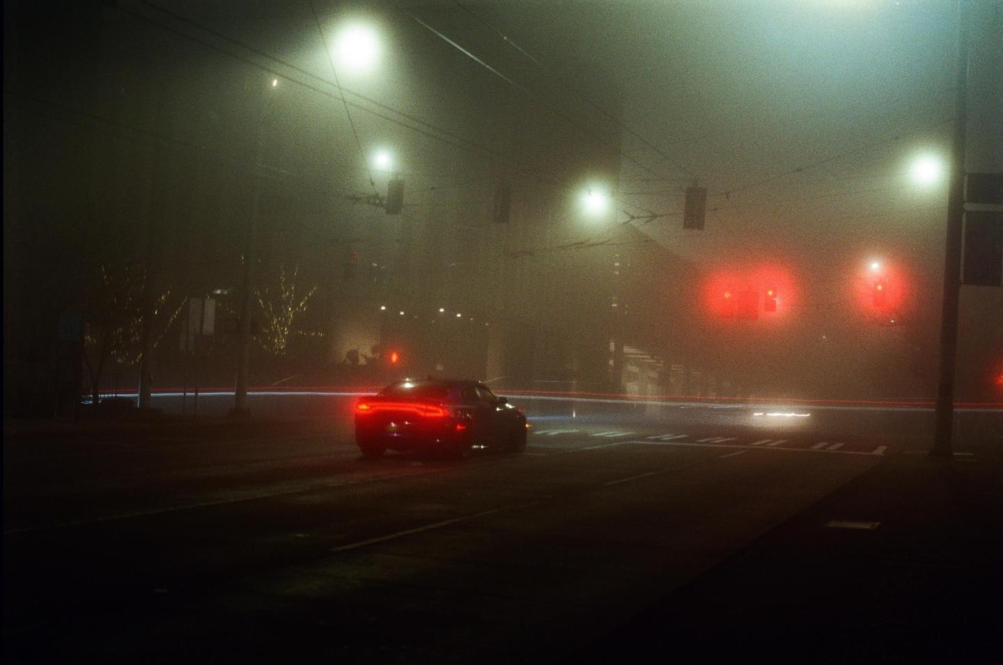 A car with illuminated taillights is visible on a foggy street at night, with streetlights and traffic lights glowing through the mist.