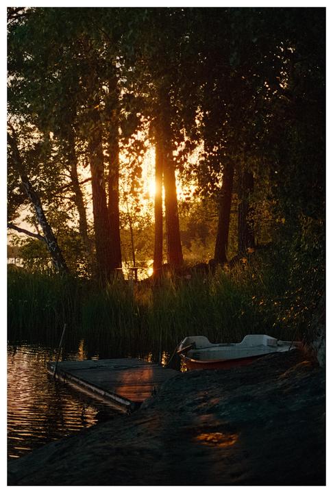 The sun sets behind trees on the shore of a lake with a small boat and a wooden dock.