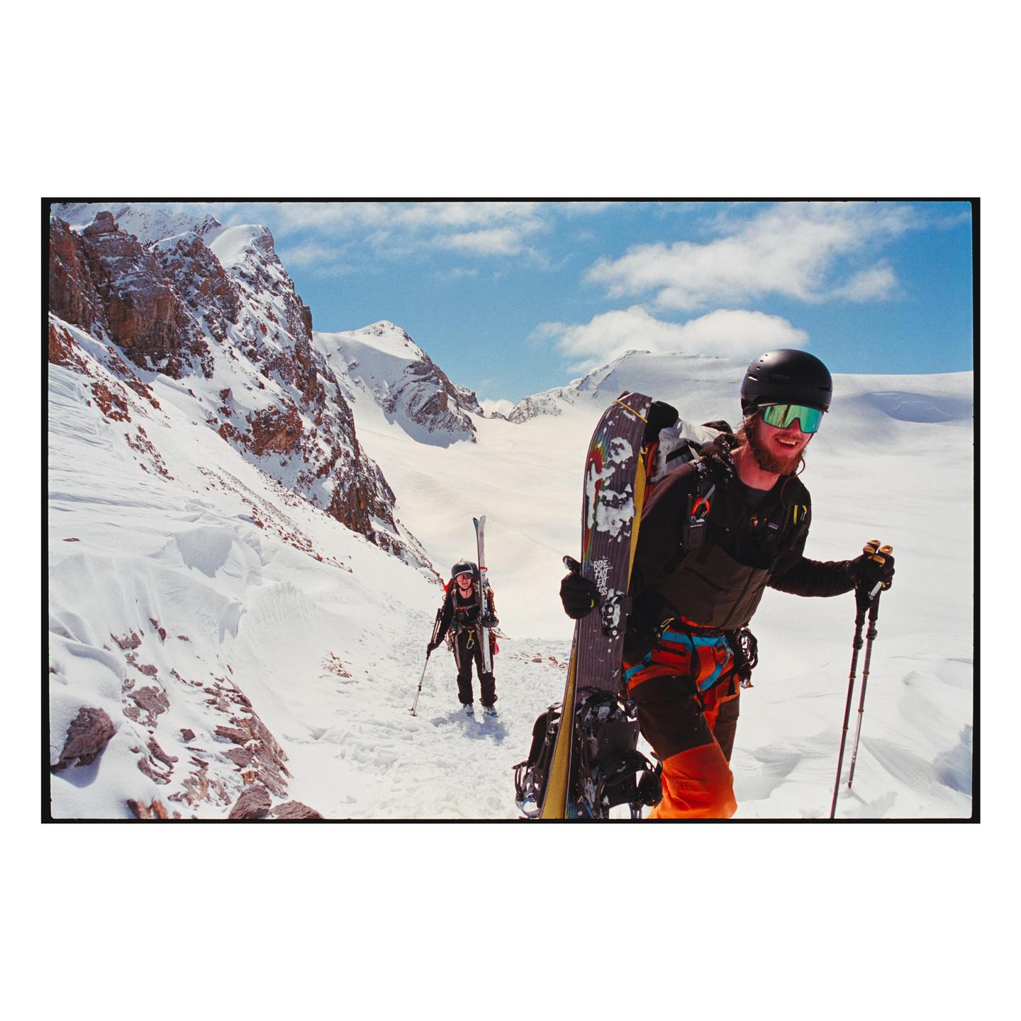 Two people are hiking up a snowy mountain path with skis and snowboards strapped to their backpacks.