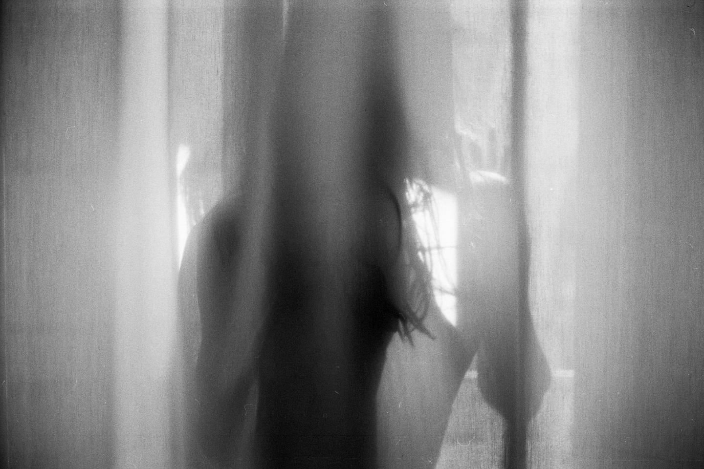 A blurry figure stands behind a curtain, with light filtering through a window.