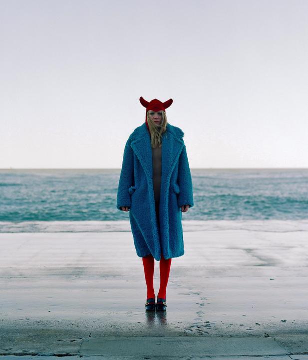 A woman in a blue coat, red tights, and a red horned hat stands on a wet concrete surface by the ocean.