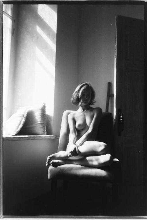 A woman sits nude on a chair in a room with light streaming through a window.