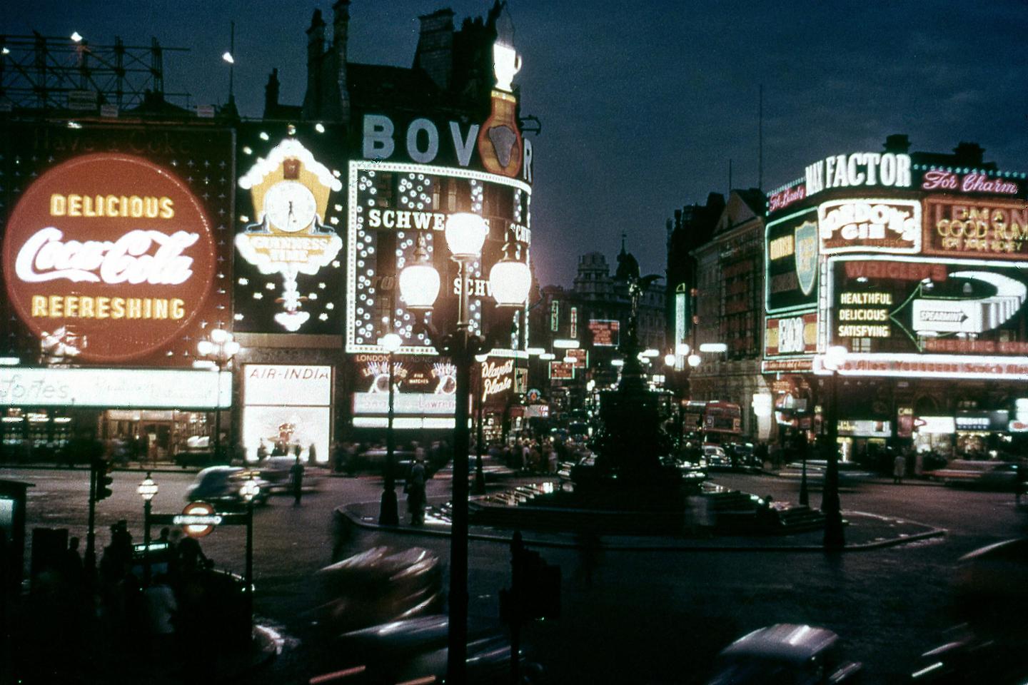A busy city street at night is illuminated by numerous neon signs and streetlights, with blurred vehicles and pedestrians indicating movement.