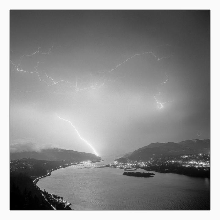 A bolt of lightning strikes a body of water surrounded by mountains and city lights at night.