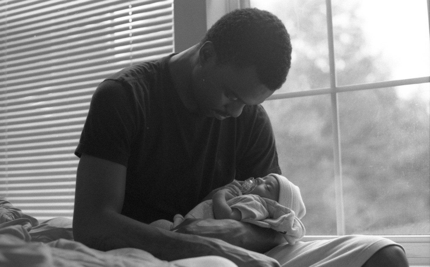 A man holds a baby with a pacifier in front of a window with blinds.