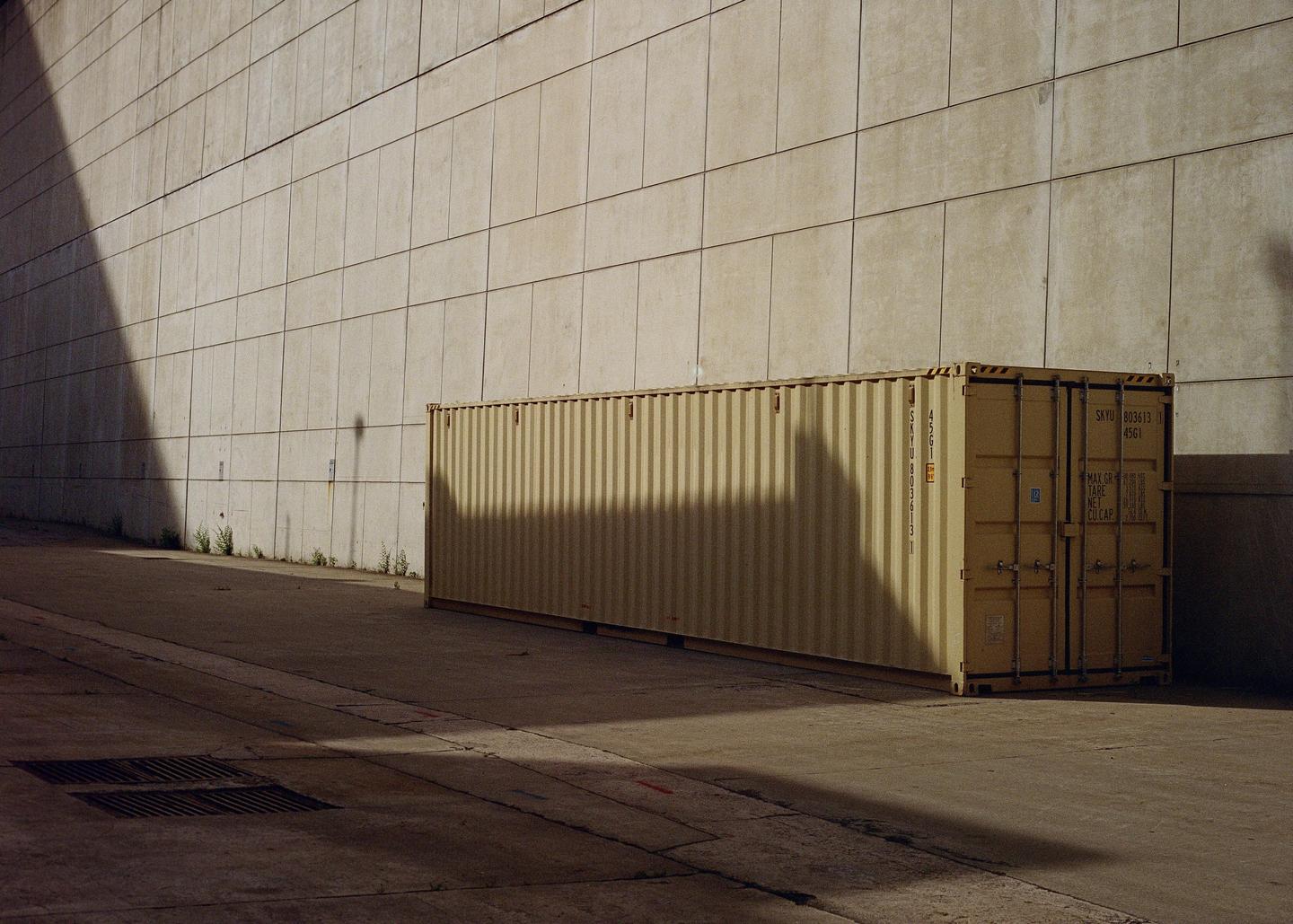 A long shipping container sits on a concrete surface next to a tall wall, with shadows cast across both.