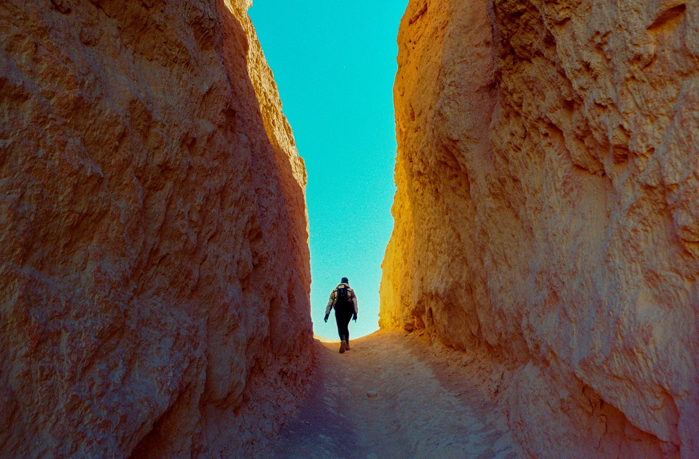 A person walks through a narrow canyon with tall rock walls under a bright blue sky.