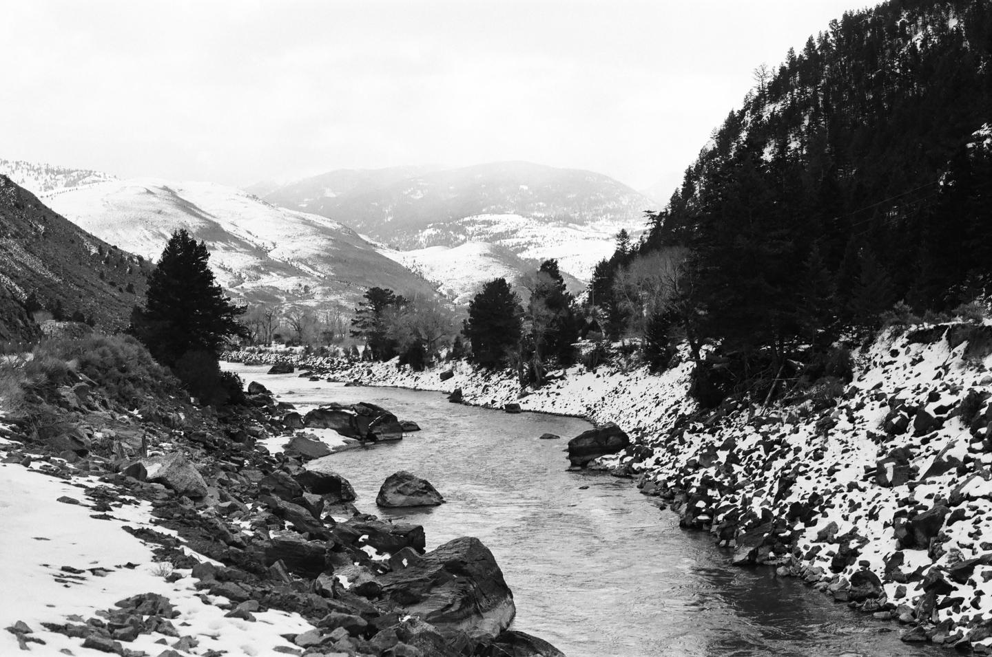 A river flows through a snowy mountain landscape with pine trees on the banks.