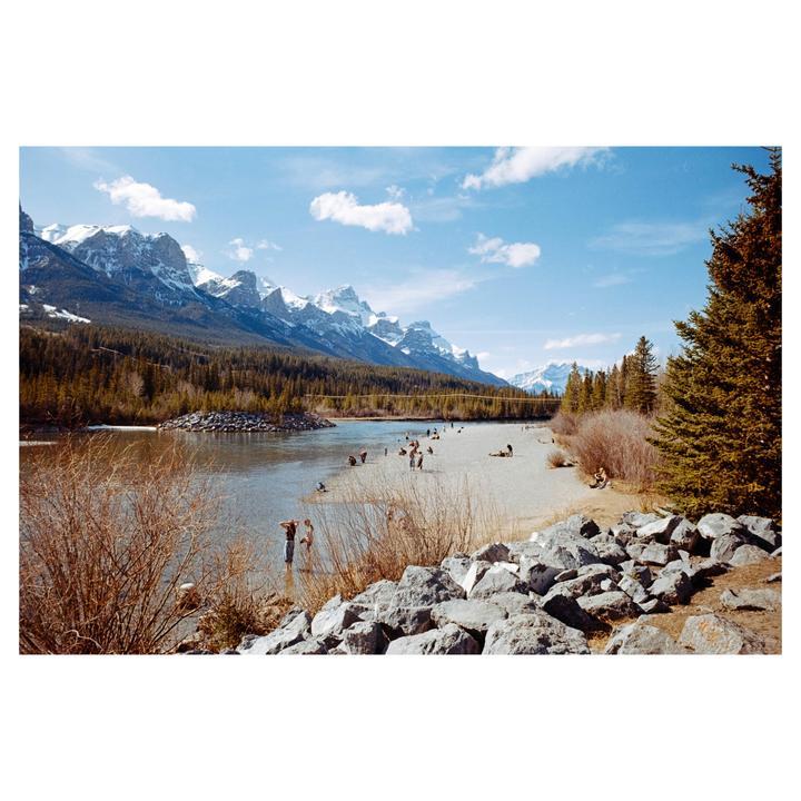 A wide river with a sandy bank is filled with people, with a forest and snow-capped mountains in the distance under a blue sky.