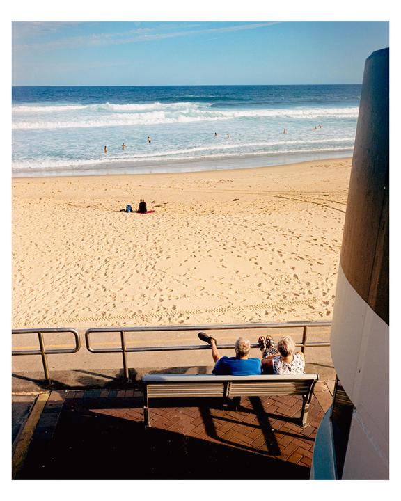 A couple sits on a bench overlooking a sandy beach with people swimming in the ocean.