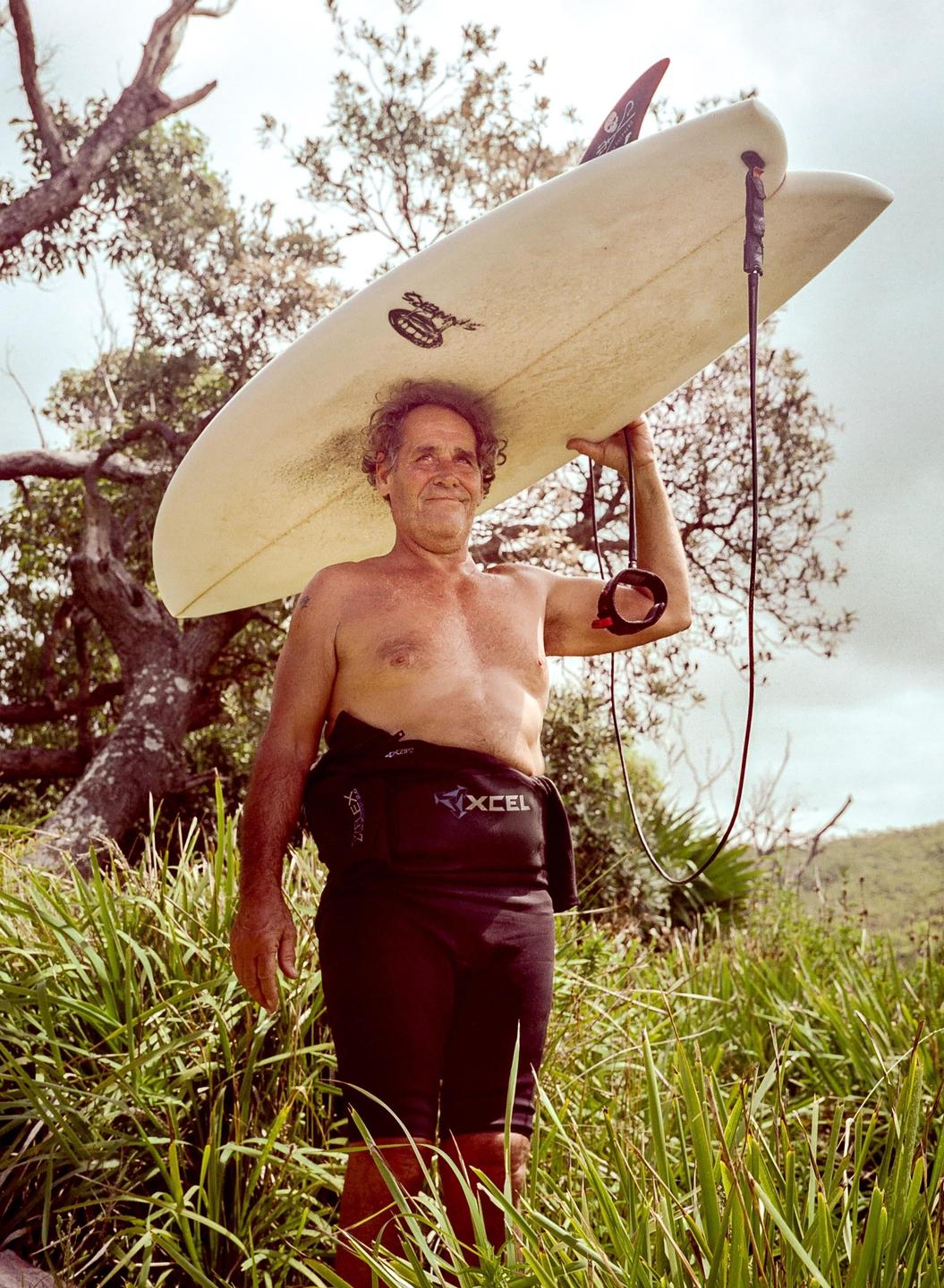 A man in a wetsuit carries a surfboard on his head through tall grass.