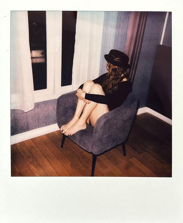 A woman wearing a hat sits in an armchair, looking out a window at dawn.