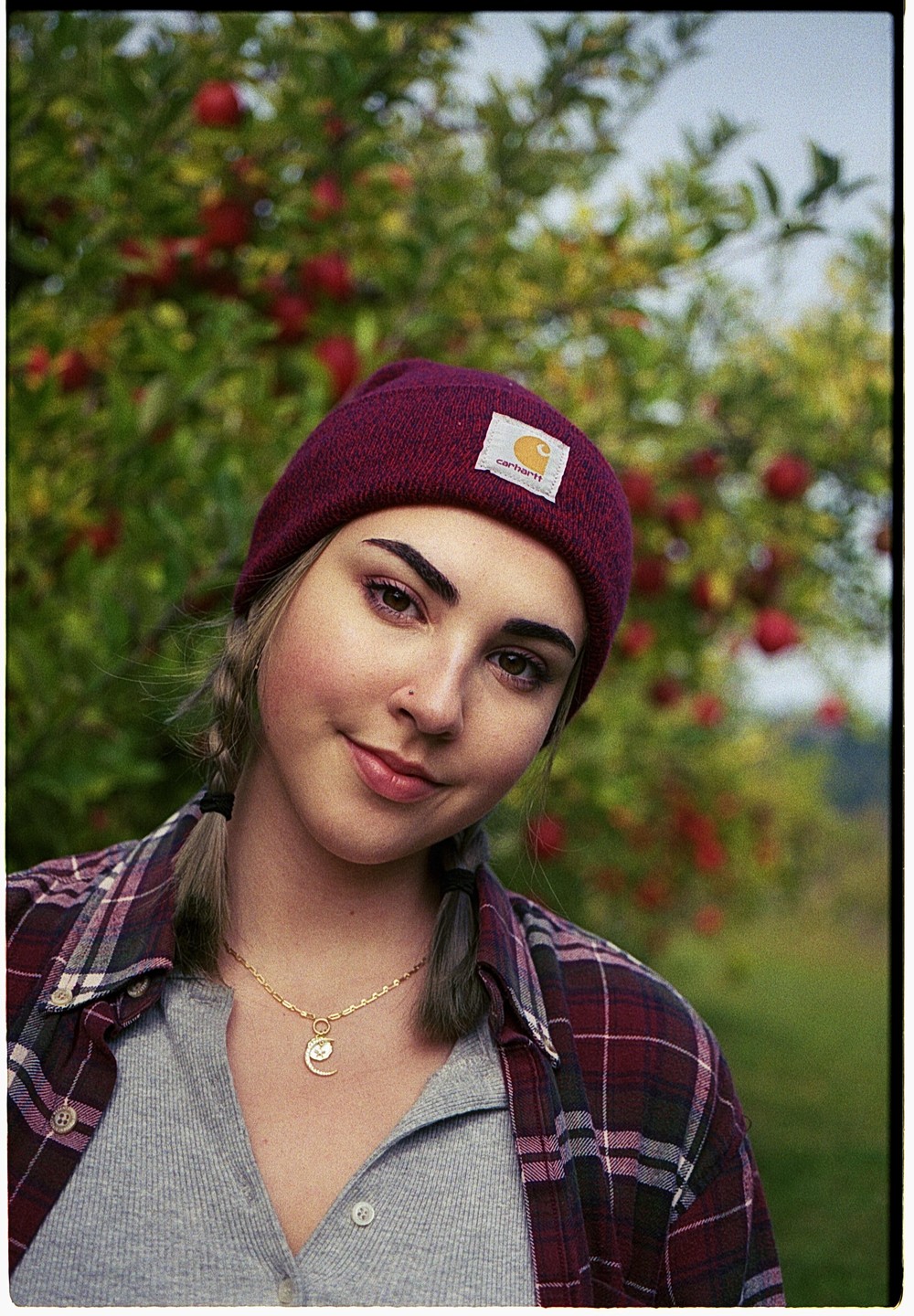 A young woman wearing a beanie and a plaid shirt smiles at the viewer, with a tree full of red apples in the background.