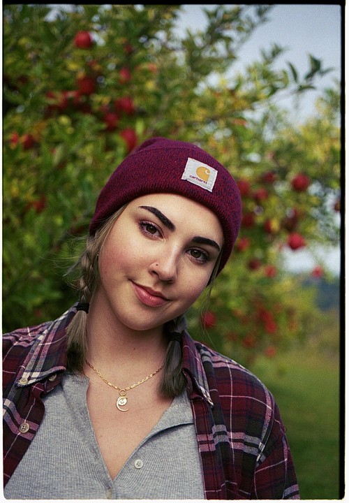 A young woman wearing a beanie and a plaid shirt smiles at the viewer, with a tree full of red apples in the background.