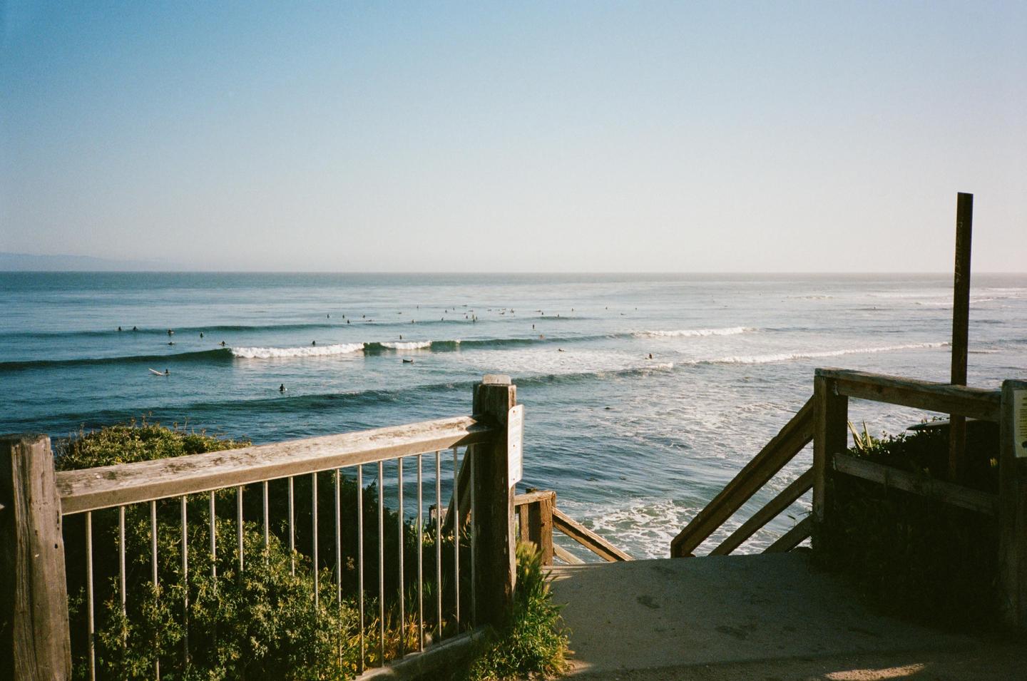A wooden railing and stairs lead down to a beach with many surfers in the water.