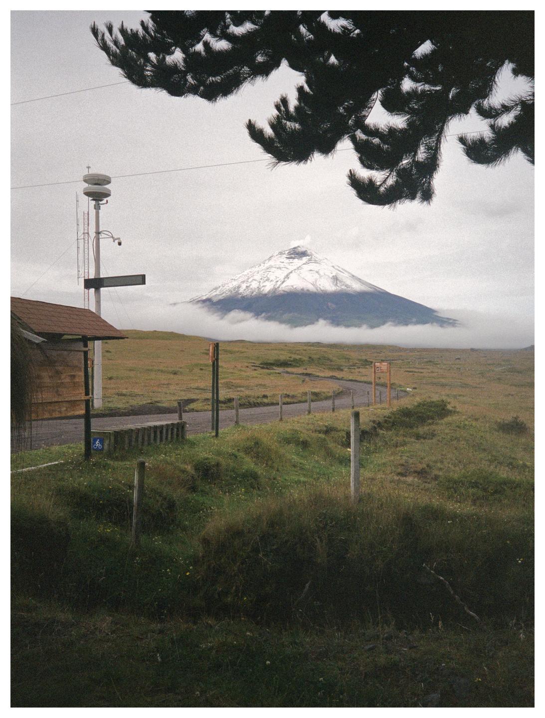 A snow-capped mountain rises above a misty landscape with a road and a small building in the foreground.