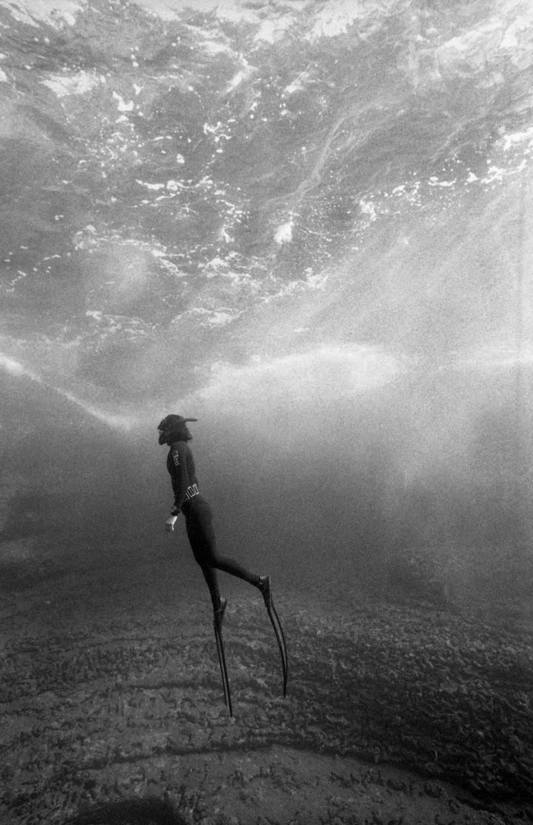 A diver in a wetsuit and fins swims underwater with light rays shining through the surface.