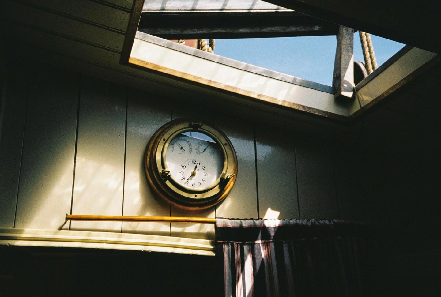 A brass clock is mounted on a wooden wall next to a striped curtain, with a skylight above revealing a blue sky.