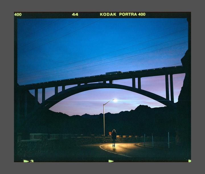 A person walks on a winding road under a large bridge at dusk, illuminated by a street light.