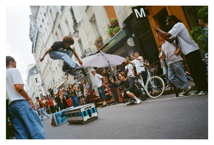 A skateboarder jumps over a model train in the street while a crowd watches.