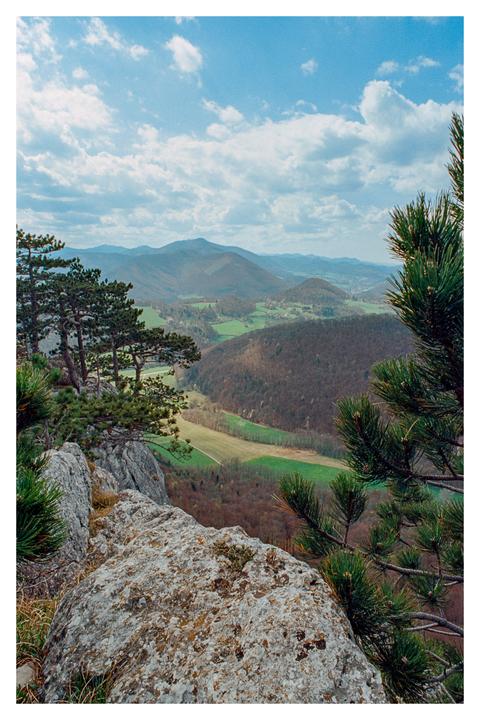 A wide view from a mountain peak shows a valley with fields and forests, and mountains in the distance under a cloudy sky.
