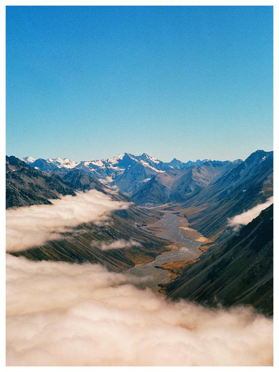 A river flows through a valley surrounded by mountains with clouds below and a clear blue sky above.