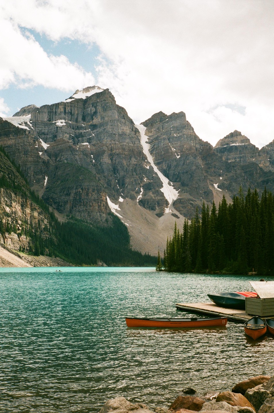 A turquoise lake with canoes and a dock is surrounded by a pine forest and snow-capped mountains under a cloudy sky.