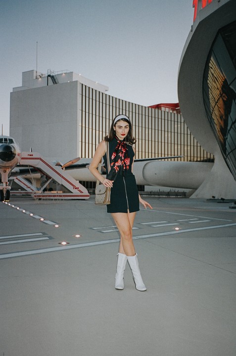 A woman in a black dress and white boots stands on an airfield with a plane and modern buildings in the background.