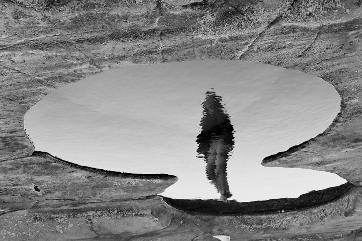 A person's reflection is visible in a puddle on a rocky surface.
