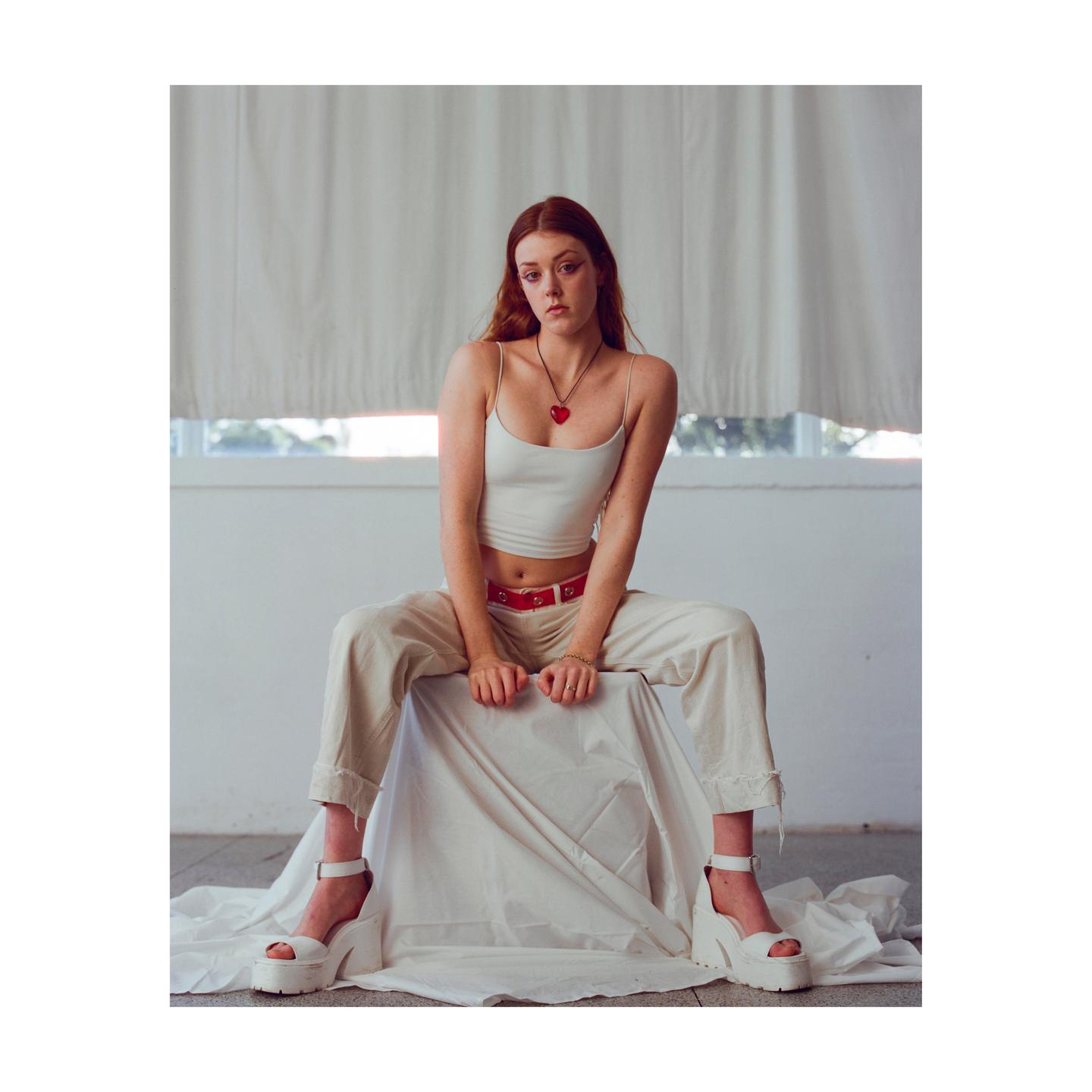 A woman with red hair sits on a draped surface, wearing a white top, light pants, and platform shoes.