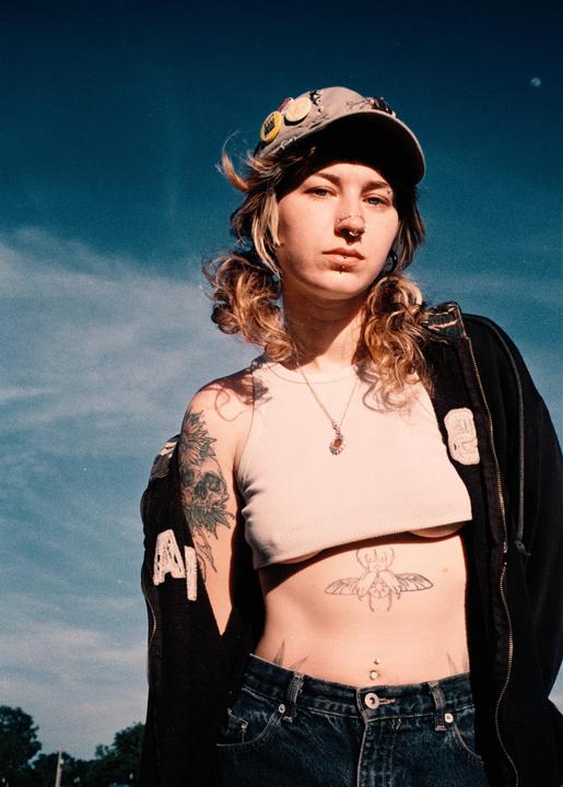 A woman with tattoos and piercings wears a cap and a cropped top against a blue sky.