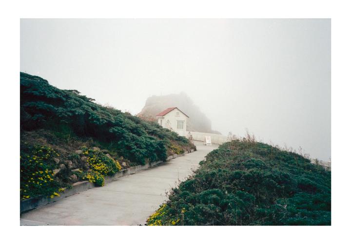 A small white house with a red roof sits on a hillside path surrounded by green bushes and yellow flowers, with a hazy sky in the background.