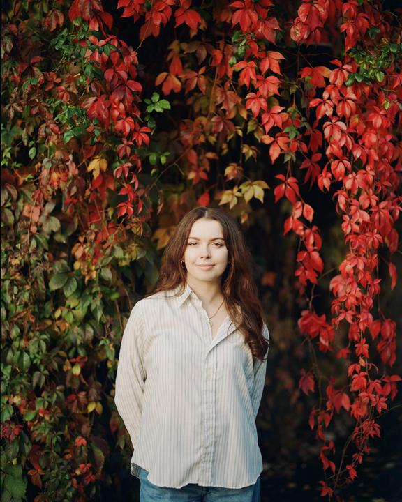 A woman stands in front of a wall of autumn foliage.