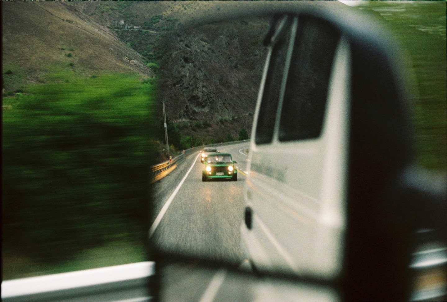 A green car with its headlights on is visible in the rearview mirror of a white van on a winding road next to a mountain.