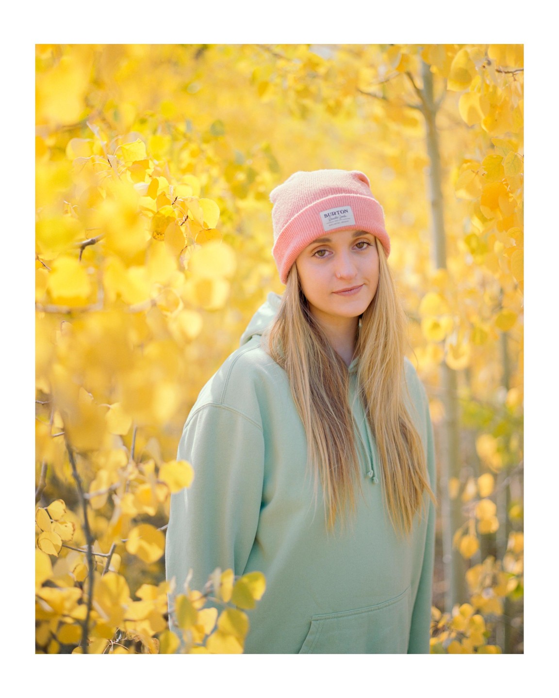 A woman wearing a pink beanie and a light green hoodie stands among bright yellow autumn leaves.