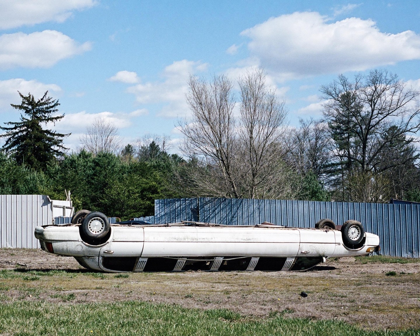 A white car is overturned in a grassy field next to a metal fence.