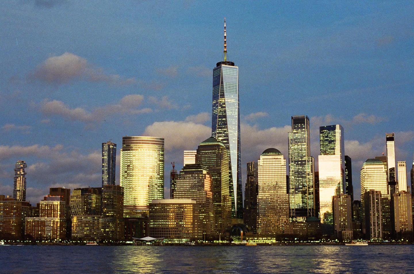 The New York City skyline is visible across the water at dusk, with the One World Trade Center skyscraper prominently featured.