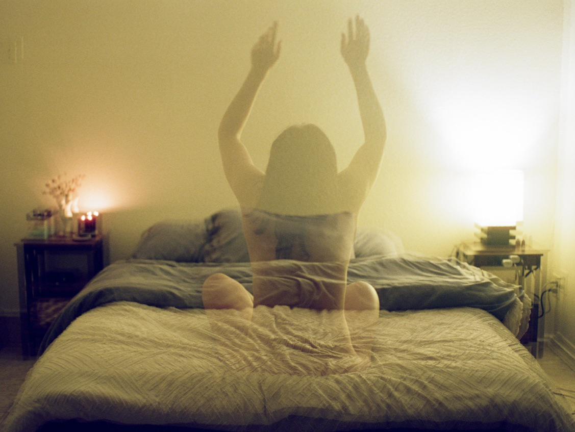 A double exposure shows a woman with her arms raised sitting on a bed in a bedroom.