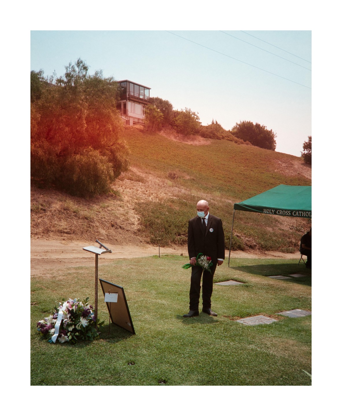 A man in a suit and face mask stands on a grassy hill next to a flower wreath and a green tent.