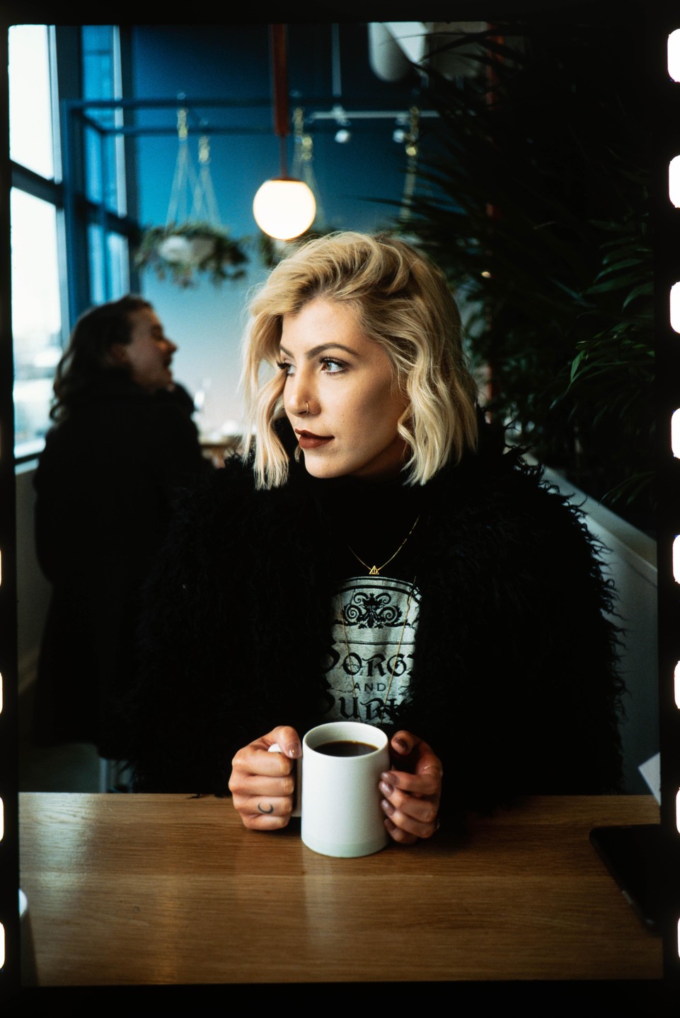 A woman with blonde hair holds a white mug at a wooden table in a cafe.