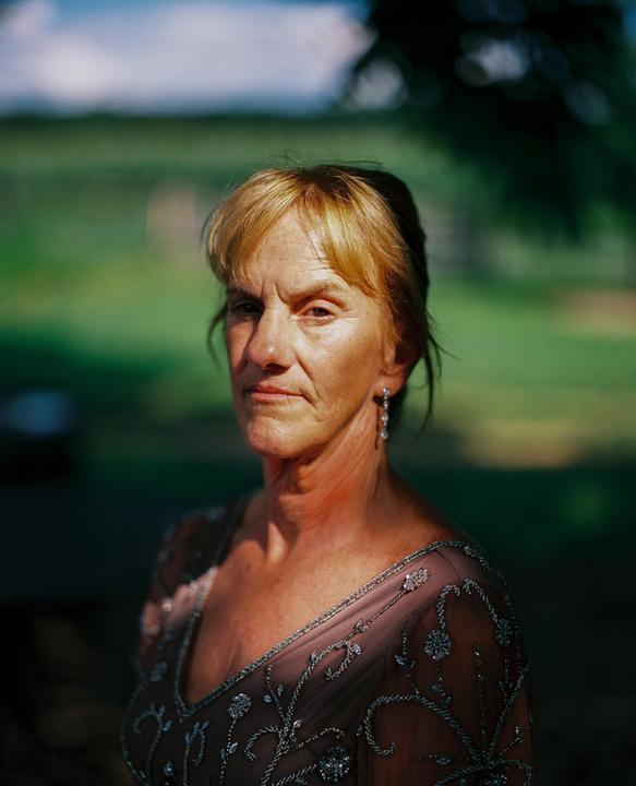 A woman with blonde hair and a beaded dress stands in dappled sunlight against a green background.